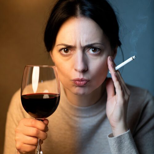 Frau wirkt nachdenklich und hält gleichzeitig ein Weinglas und eine Zigarette – Symbol für den inneren Konflikt beim gleichzeitigen Aufhören von Alkohol und Rauchen
