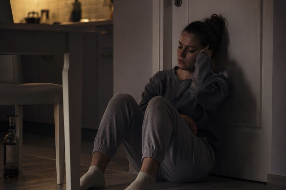 Frau sitzt nachts erschöpft auf dem Küchenboden vor einer geschlossenen Tür. Die Wohnung ist dunkel, nur warmes Licht aus der Küche fällt in den Raum. Neben ihr liegt ein Handy, unter dem Tisch steht halb verborgen eine Flasche Alkohol. Stimmung von Einsamkeit, innerem Kampf und dem Wunsch nach einem Ausweg.