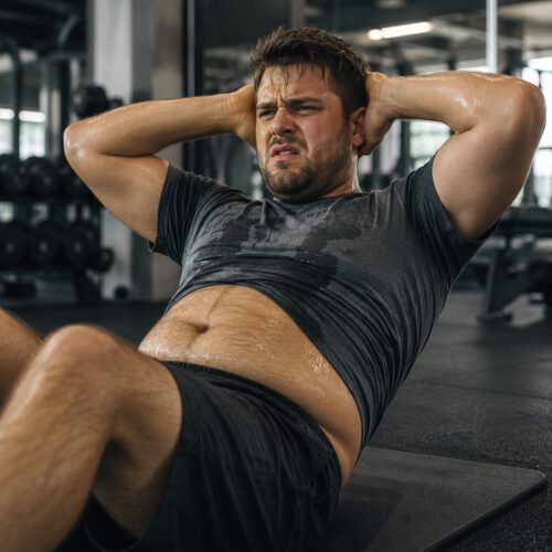 Junger Mann mit sichtbarem Bierbauch macht im Fitnessstudio erschöpft Sit-ups. Sein T-Shirt ist schweißnass, sein Gesicht zeigt Anstrengung und leichten Frust. Das Bild verdeutlicht, dass Sit-ups zwar die Bauchmuskeln stärken, das gesundheitlich besonders problematische viszerale Bauchfett jedoch nicht gezielt abbauen – insbesondere dann nicht, wenn regelmäßiger Alkoholkonsum die Fettverbrennung zusätzlich hemmt.